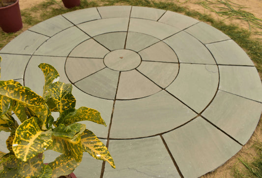 A round outdoor stone paver patio with concentric circular and radial line patterns, shown on a sandy ground with some green grass and a potted variegated plant in the foreground.